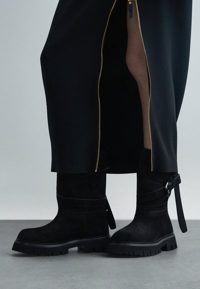 Μαύρες σουέντ μπότες μέχρι το γόνατο με χοντρές σόλες φορεμένες με μαύρη φούστα που διαθέτει χρυσό φερμουάρ στην πλευρά αποκαλύπτοντας το πόδι.