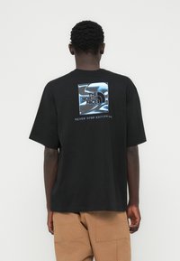 Μαύρο βαμβακερό μπλουζάκι με κοντά μανίκια, με γραφικό λογότυπο σε μπλε και λευκό στην πλάτη. Το κείμενο κάτω από το λογότυπο γράφει «NEVER STOP EXPLORING».