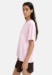 Jasnoróżowa koszulka z krótkim rękawem i okrągłym dekoltem, wykonana z bawełny. Posiada małą czerwoną metkę na dolnym brzegu. Model nosi ciemne szorty.