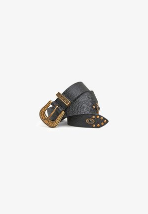 Ceinture en cuir noir texturé avec boucle dorée ornée et clous dorés, certains en forme d'yeux, enroulée sur un fond blanc.