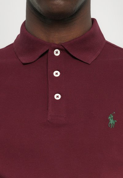 Bordo polo majica s klasičnim ovratnikom, placketom na dva dugmeta i izvezenim zelenim logotipom. Napravljena od teksturiranog pamučnog materijala.