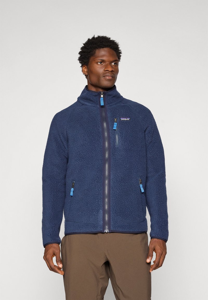 Navy fleece jacket with a high collar, zip closure, and contrasting blue zipper pulls. Features two side pockets and a logo on the chest.