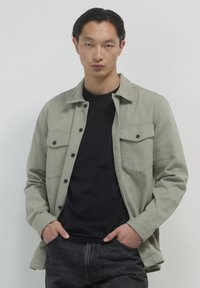 Camicia button-up verde chiaro con due tasche sul petto, bottoni neri e vestibilità rilassata, indossata sopra una maglietta nera e jeans scuri.