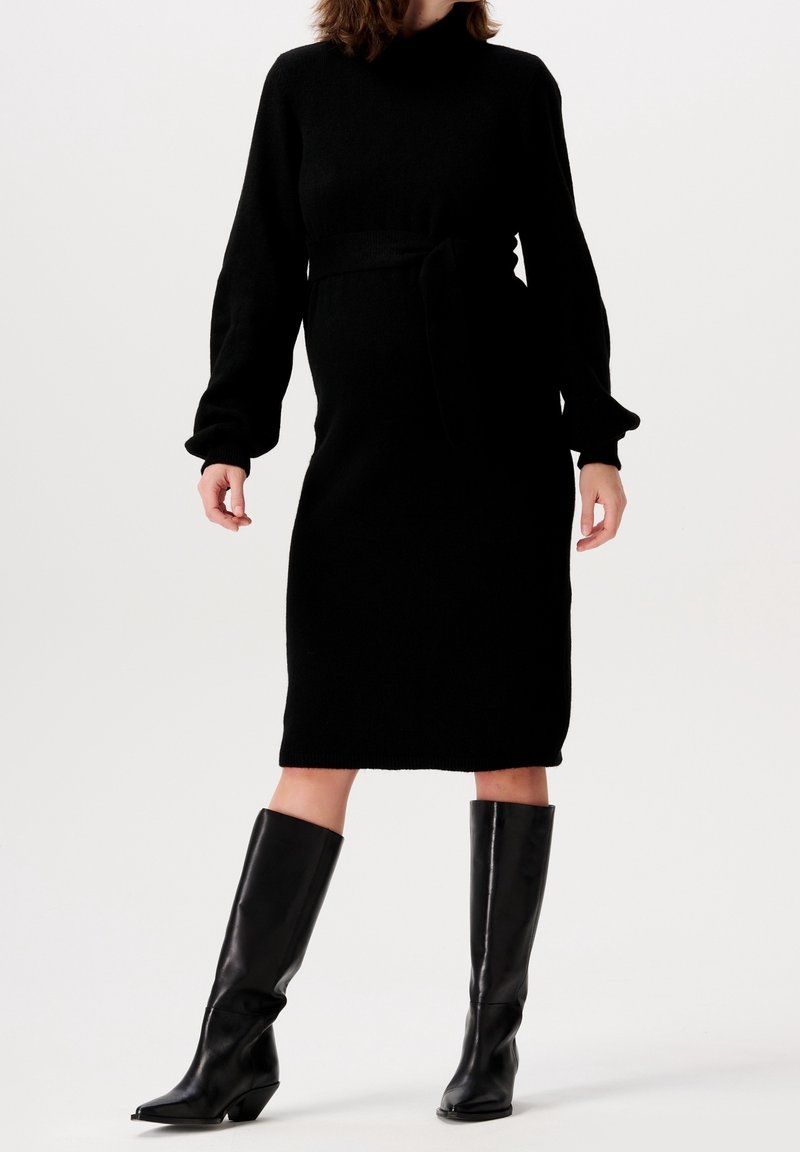 Femme portant une robe midi noire à manches longues avec une ceinture à la taille et des bottes en cuir noires montant jusqu'aux genoux sur un fond blanc.