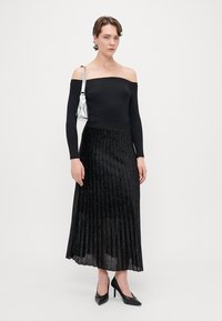 Sort plisseret nederdel med glitrende accenter, parret med en off-shoulder sort top. Modellen holder en skinnende sølv taske og har højhælede sko på.