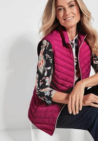 Fuchsia gewatteerde vest met een hoge hals, voorzien van een rits aan de voorkant en zijzakken, gedragen over een bloemetjesblouse met driekwartmouwen.
