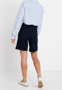 Tmavě modré šortky s ohrnutými lemy, které mají dvě zadní kapsy. Kombinovány s světle modrou košilí s dlouhým rukávem a bílými espadrilkami.