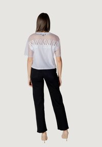 Camiseta blanca corta con mangas de malla transparente y patrón de triángulo bordado en la parte posterior, combinada con pantalones negros. Tacones visibles.