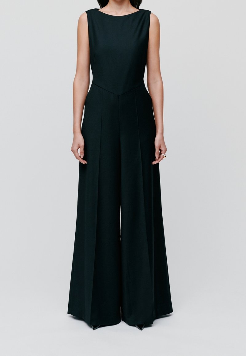 Femme portant une combinaison noire sans manches à jambes larges avec un corsage ajusté, debout devant un fond uni gris clair.