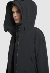 Parka noire avec une grande capuche, fermeture éclair à l'avant et poches latérales. Fabriquée en tissu lisse avec des détails de couture subtils.
