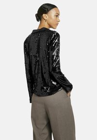 Schwarze Paillettenbluse mit Kragen, langen Ärmeln und lockerer Passform; kombiniert mit hellgrauen Hosen. Glatte, reflektierende Textur und Glanz.