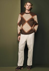Brown, beige, and rust diamond-patterned sweater, cream trousers, and dark brown shoes with light-colored soles. Simple, textured design.