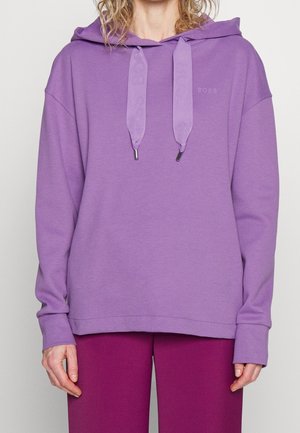 Personne portant un sweat à capuche violet clair avec de larges cordons et un pantalon violet foncé, debout devant un fond uni.