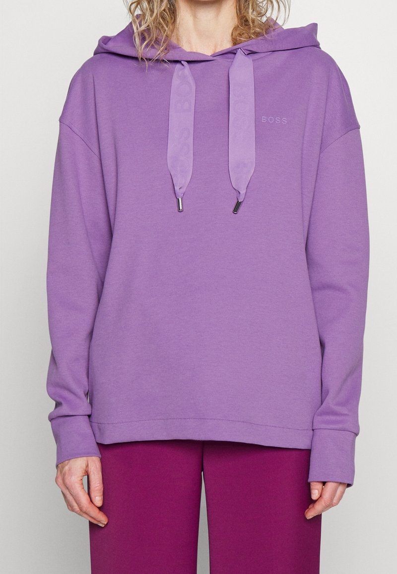 Personne portant un sweat à capuche violet clair avec de larges cordons et un pantalon violet foncé, debout devant un fond uni.