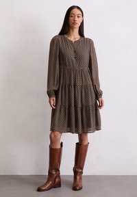 Robe marron à motifs avec un décolleté boutonné, des manches longues et un ourlet à volants. Assortie à des bottes en cuir marron hautes. Texture lisse.