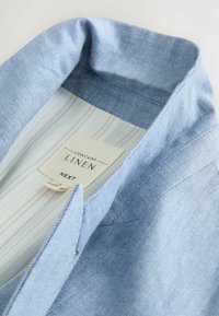 Světle modrá lněná košile s límcem, texturovaným povrchem a pruhovanou vnitřní podšívkou. Etiketa uvádí "Obsahuje len" a "NEXT."