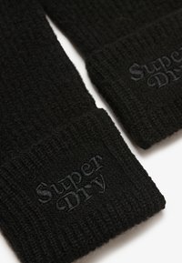 Guanti in maglia nera con polsini a coste e superficie testurizzata. Il logo "Superdry" è ricamato in nero coordinato sul polsino.