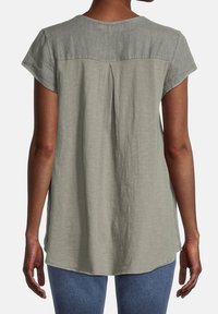 Haut gris clair à manches courtes avec un panneau supérieur légèrement texturé, détail de dos plissé et un ourlet incurvé, porté avec un jean bleu.