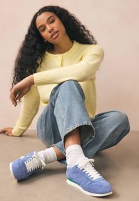 Sneakers in camoscio blu con lacci bianchi e dettagli verdi, abbinate a calzini bianchi. Il modello indossa un maglione giallo e jeans azzurri.