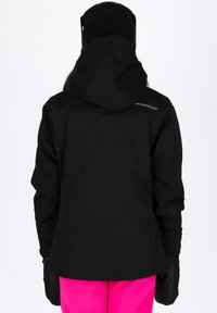 Giacca nera impermeabile con capuccio, caratterizzata da una texture liscia e da un logo sulla parte superiore della schiena, abbinata a pantaloni rosa e guanti neri.