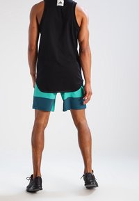 Svart ärmlös tanktopp med rundad nederkant. Teal och mörkgröna shorts med en tvåfärgad design, kombinerat med svarta löparskor.