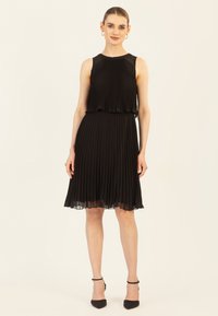 Schwarzes, plissiertes Kleid mit ärmellosem Design, geschichteter Oberseite und transparenter Säume. Getragen mit schwarzen Ankle-Strap-Absätzen und Kreolen.