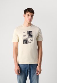 Young man wearing a beige short-sleeve T-shirt with abstract black and white graphic print, paired with blue jeans, standing against a plain background.