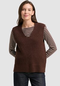 Frau mit braunem Haar, die eine braune gestrickte ärmellose Weste über einem braun-weißen gestreiften Langarmshirt und dunklen Jeans trägt.