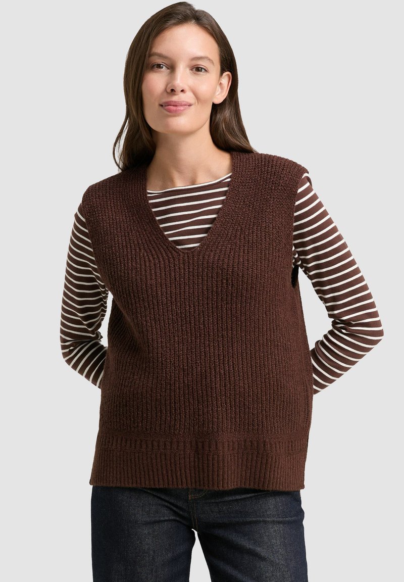Frau mit braunem Haar, die eine braune gestrickte ärmellose Weste über einem braun-weißen gestreiften Langarmshirt und dunklen Jeans trägt.