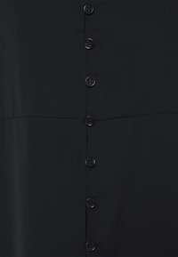 Tissu noir avec une rangée verticale de sept boutons ronds noirs à quatre trous chacun, cousus le long de la couture centrale.