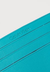Portofel turcoaz cu suprafață texturată, având striații orizontale și logo-ul embosat "Calvin Klein". Design compact și dreptunghiular.