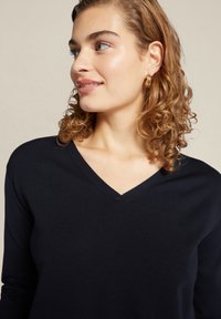 Marineblauwe v-hals sweater van zacht breimateriaal, met lange mouwen en een ontspannen pasvorm. Krullend haar en oorringen maken de look compleet.
