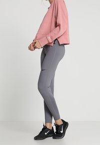 Femme portant un sweat-shirt rose, un legging Nike gris et des baskets Nike noires, debout de profil sur un fond clair uni.