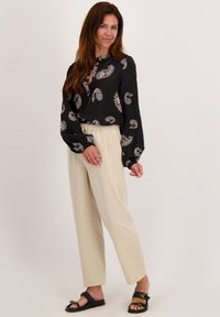 Zwarte blouse met lange mouwen en een wit paisleypatroon, gecombineerd met beige gestreepte pantalons en zwarte sandalen met een gouden accent gesp.