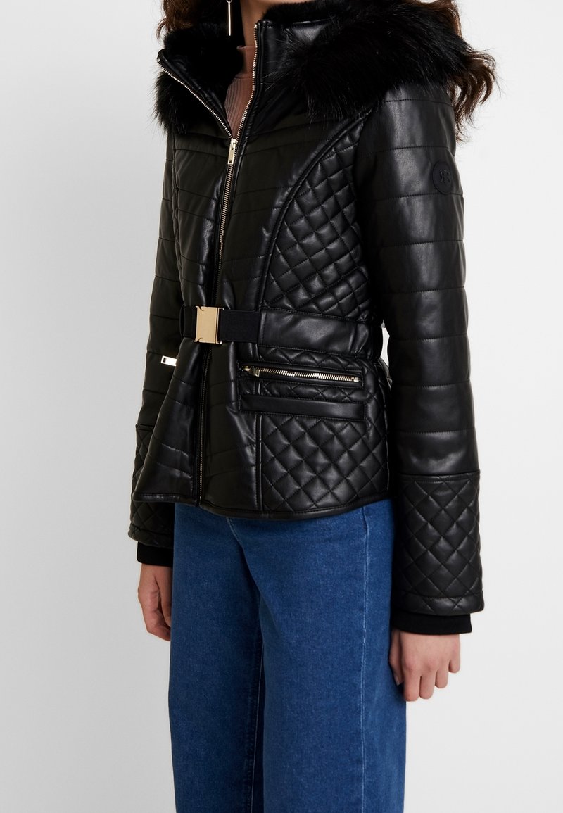 Veste en cuir matelassé noir avec un col en fausse fourrure, accessoires en métal doré, taille ceinturée et poches zippées, associée à un pantalon en denim bleu.