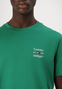 Zöld pamut póló, kerek nyakkal, rövid ujjakkal, a mellkason "Tommy Hilfiger" felirattal és a megalakulás évével ellátott logóval.