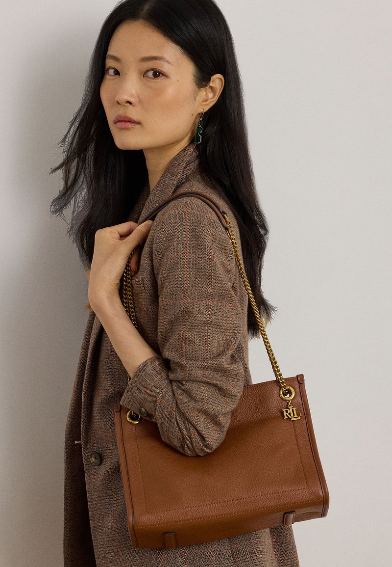 Femme aux longs cheveux noirs porte un blazer marron à carreaux et porte un sac à main en cuir marron avec une bandoulière en chaîne dorée et un charm RL sur l'épaule.