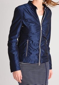 Navy blue nylon jacket with a fitted design, collar, side pockets, and silver zipper accents. Worn with a striped skirt.