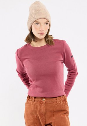 Jonge vrouw met een beige gebreide muts, een roze longsleeve met schouderknopen en roestkleurige broek, poserend met handen achter de rug.
