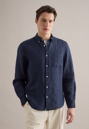 Hombre joven con una camisa azul marino de manga larga abotonada con rayas finas blancas y pantalones beige, de pie frente a un fondo liso.