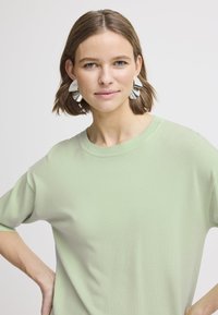 Maglione verde chiaro con scollo rotondo e maniche corte, caratterizzato da una consistenza morbida. Sono indossati grandi orecchini geometrici in argento.