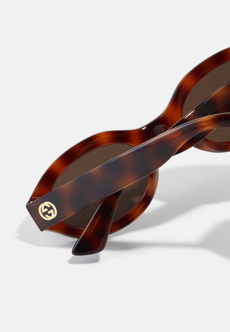 Schildpatt-Sonnenbrille mit runden Gläsern. Kühner, glänzender Rahmen mit sanften Kurven. Die Bügel sind mit einem goldenen Logo-Akzent und einer glatten Textur versehen.