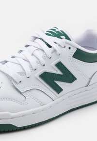 Bílé tenisky s zelenými akcenty, s koženým svrškem, šněrováním a strukturovanou podrážkou. Výrazné zelené logo "N" na boku.