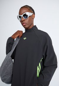 Black jacket with high collar and green Adidas logo, paired with gray shoulder bag. Model wears white cat-eye sunglasses, featuring a sleek design.