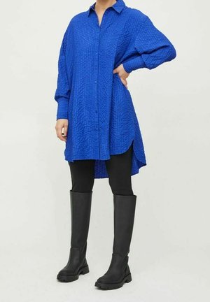 Osoba ubrana w długą, niebieską, teksturowaną koszulę na guziki, czarne legginsy oraz czarne, masywne kozaki za kolano, stojąca z jedną ręką na biodrze.