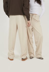 Pantalons beiges ajustés avec une coupe décontractée, comportant des poches avant plates et des détails de pli subtils. Un des porteurs porte des chaussures à motifs, l'autre des baskets.