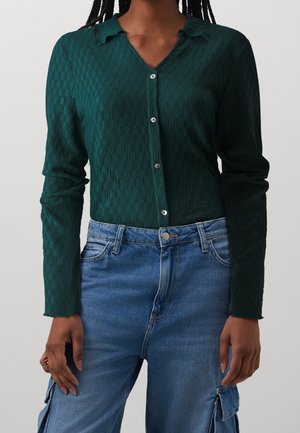 Frau trägt ein dunkelgrünes, strukturiertes, langärmeliges Hemd mit Knöpfen und blaue, hoch taillierte Cargo-Jeans mit Taschen.