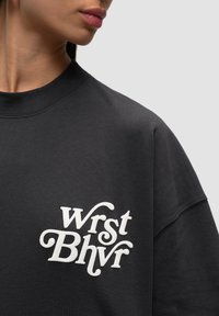 Sweatshirt noir avec un col rond, arborant le texte brodé en blanc "Wrst Bhvr." Texture de tissu douce, coupe décontractée et manches courtes.