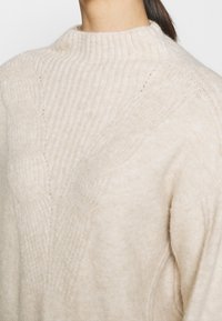 Pull en tricot beige avec un col montant côtelé, des motifs texturés et une coupe ample, avec des détails de couture visibles.
