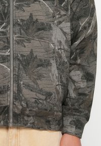 Kapucňová zipsová bunda v tmavosivej farbe s motívom listov. Textúrovaný povrch s kontrastnými svetlými konárikmi a elastickým lemom.